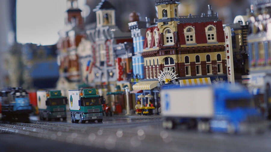 A closeup image of cityscape made of lego. Street scene includes buildings, street, signs, semi-trailer trucks and train on train tracks.