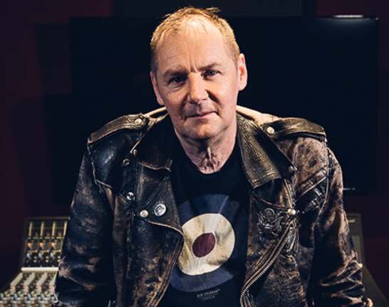 Man wearing leather jacket, leaning on sound board, looking at the camera.