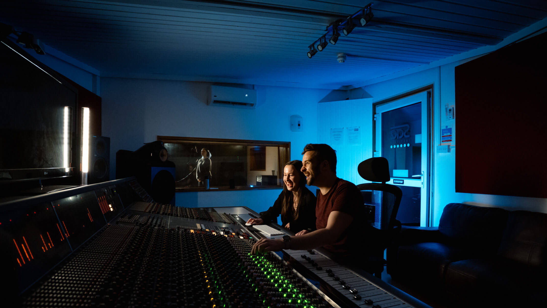 Two people laughing while recording live music. A person is in the background in a sound studio with headphones on in front of a microphone. The people appear to be in front of a backlit audio mixing console with sliders and buttons.