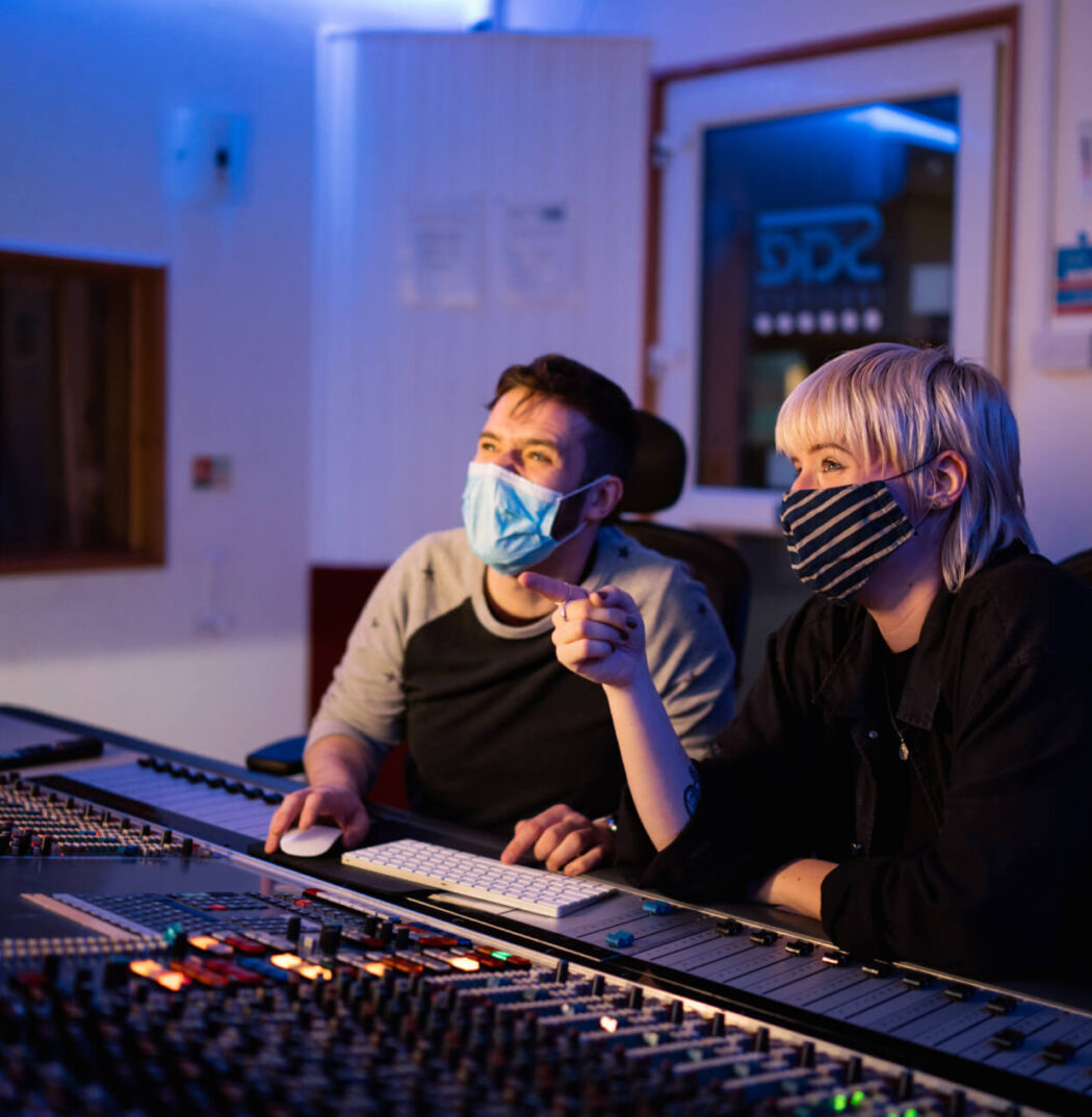 Two music students wearing face masks sit in an audio studio. One is moving a mouse and using a keyboard. The other is pointing off camera.