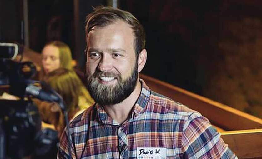 Man stands in lecture with camera in the background. Sticker on chest reads David K.