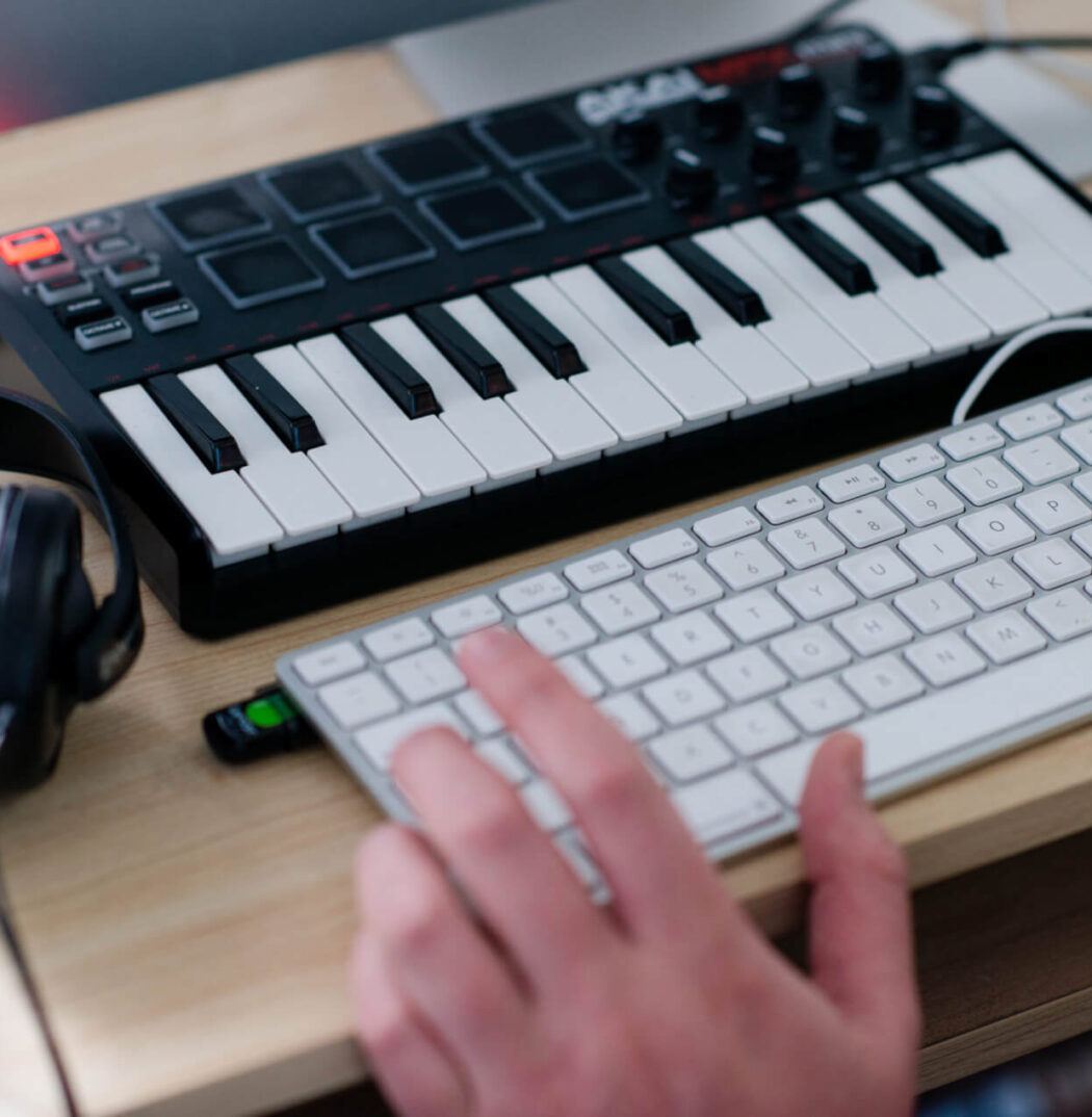 Image of audio keyboard, computer keyboard and headphones on desk. Hand hovers over QWERTY keyboard.