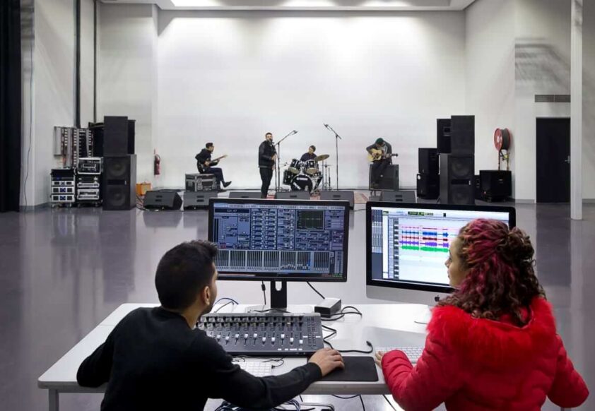 SAE audio students recording live 4-piece band on a soundstage. Two people sit at monitors in foreground. Band in backdrop.