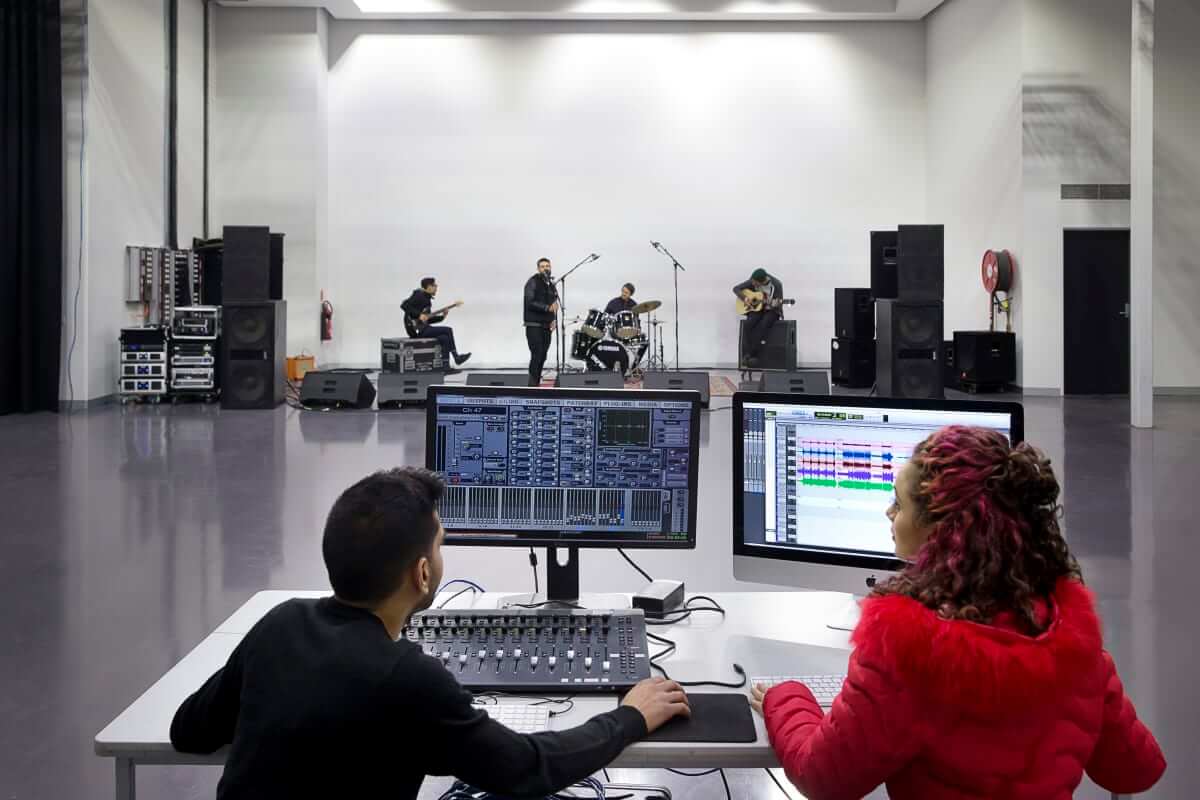 SAE audio students recording live 4-piece band on a soundstage. Two people sit at monitors in foreground. Band in backdrop.