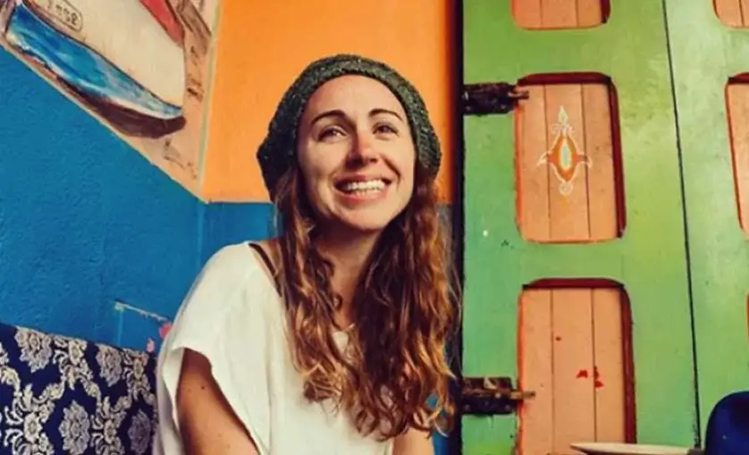 Woman in a colourful room smiles to camera. She has long hair and is wearing a beanie.