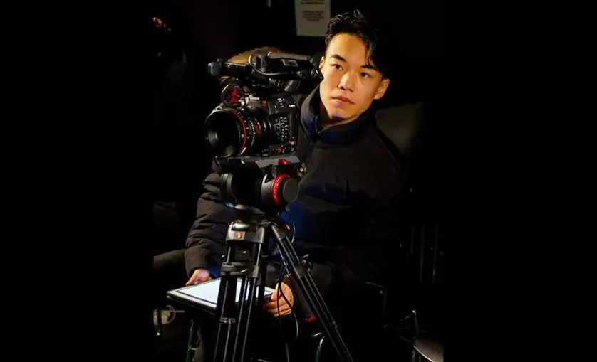 Man crouches behind a professional camera on a tripod, looking through viewfinder and holding an iPad.