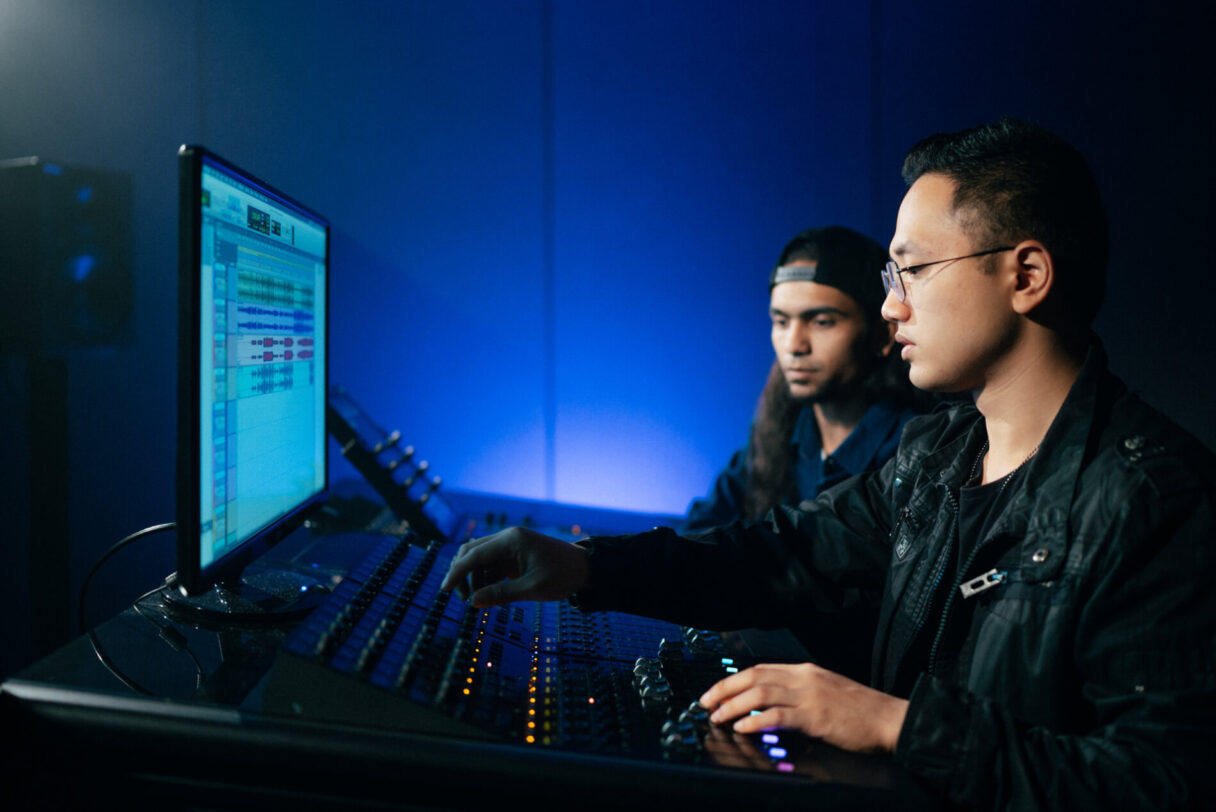 Two people sitting in front of and using Audio mixing Desk and monitor with editing software on screen. The room is backlit blue but dark.