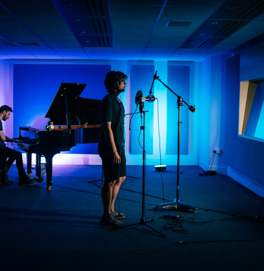 Two people in a recording studio. One male is seated at a grand piano, both hands and feet in contact with the piano. The other man is standing in front of two microphones on stands (with a shield). The room is back lit blue.