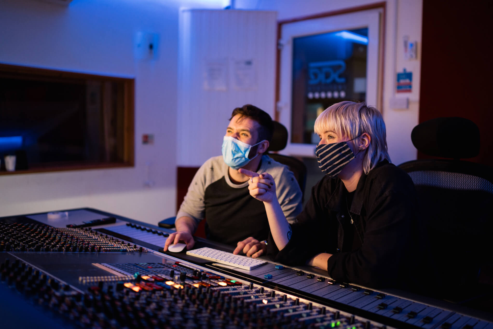 Two music students wearing face masks sit in an audio studio. One is moving a mouse and using a keyboard. The other is pointing off camera.