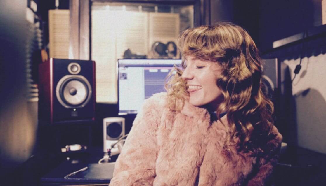 Person sitting in studio, smiling. Wearing faux fur coat