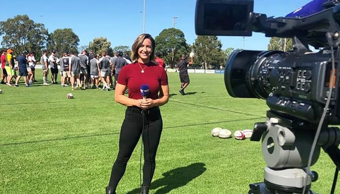 Female TV presenter standing with microphone on rugby field.