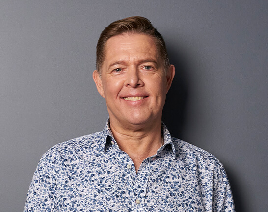 Dean Pearson. Portrait headshot of man smiling, wearing a blue printed shirt.