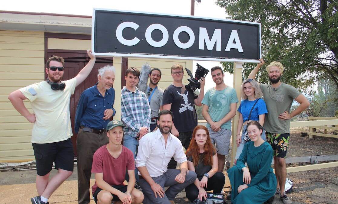 Film crew of many people - actors, camera operator and sound people stand under 'Cooma' sign