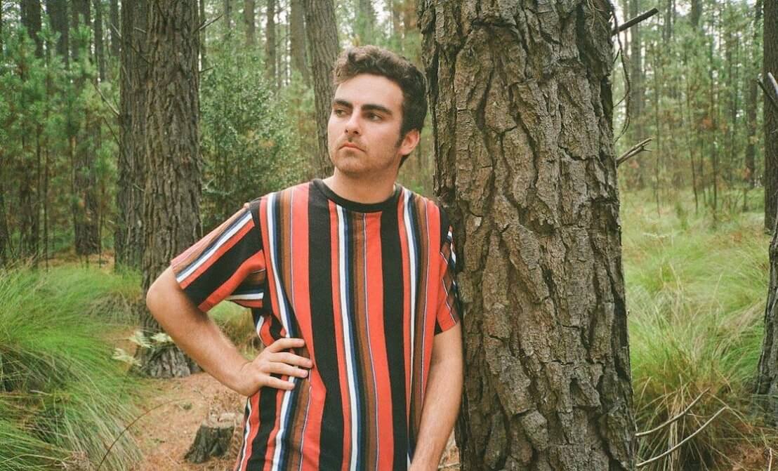 Man in striped tshirt leaning on a tree.
