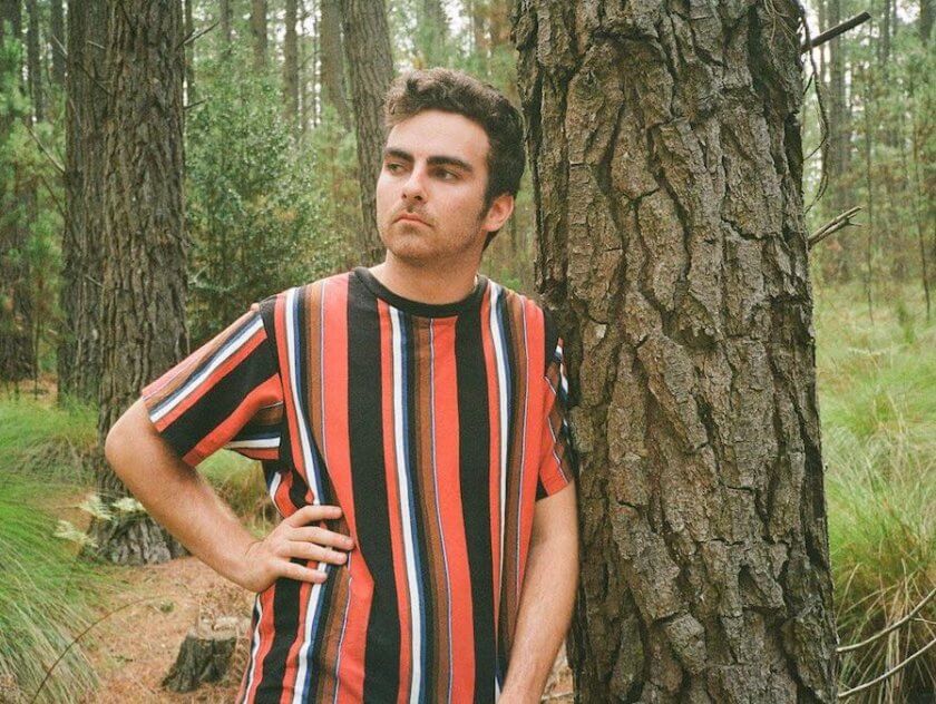 Man in striped tshirt leaning on a tree.