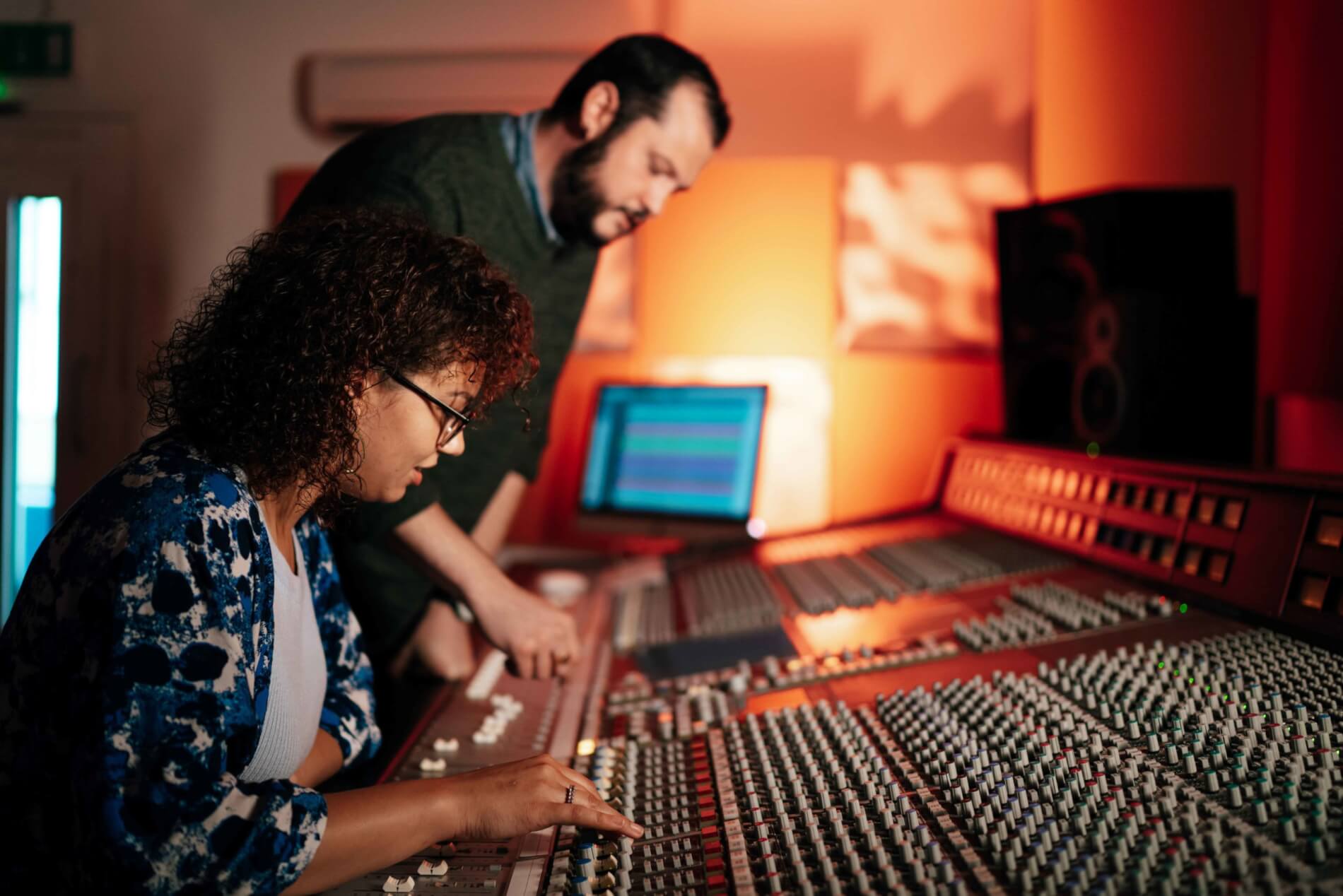 Girl sits at audio mixing analgoue desk with man standing. Both are adjusting levels and knobs.
