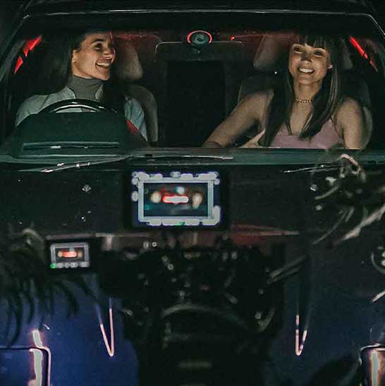 Two women are seated in front seat of car. A camera floats in front of wind shield with cables and monitors and lighting.