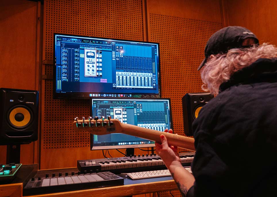 A man with his back turned to camera playing an electric guitar, with various electronic music instruments and two monitors displaying production software in front of him