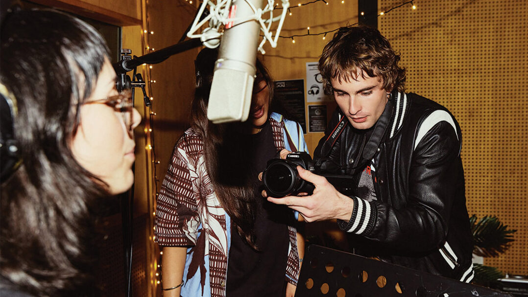 A young male student filming a female music student singing in to a boom microphone, with another female student watching from behind the camera - Create for a day at SAE