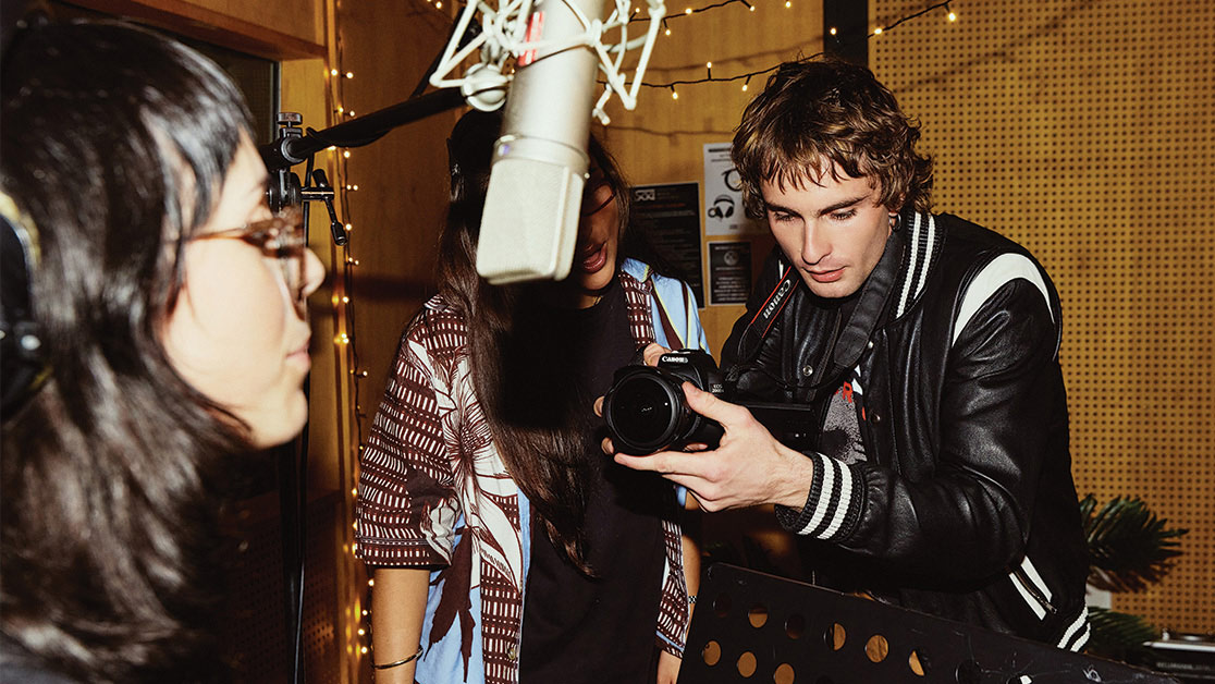 A young male student filming a female music student singing in to a boom microphone, with another female student watching from behind the camera - Create for a day at SAE