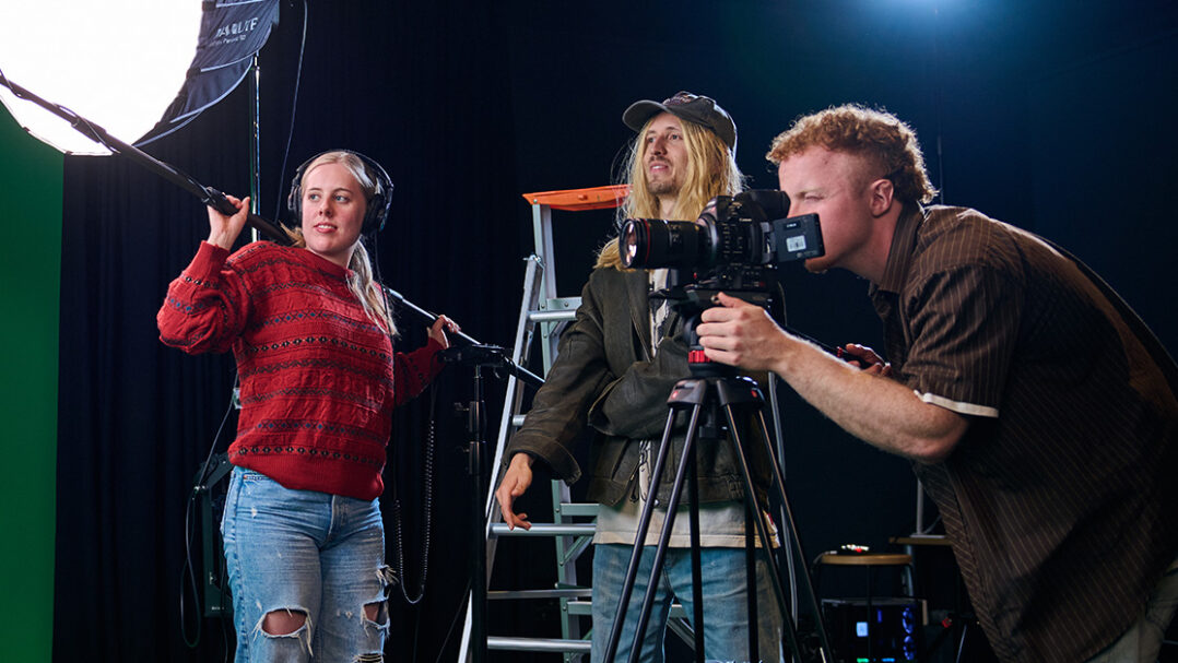 Three SAE University College film students standing together, with one operating a camera and another holding a boom microphone