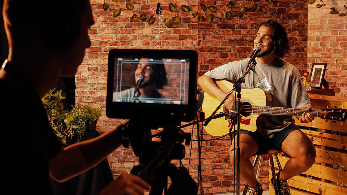 A cameraman and monitor captures a musician - sitting on a stool in front of microphone while singing and playing an acoustic guitar