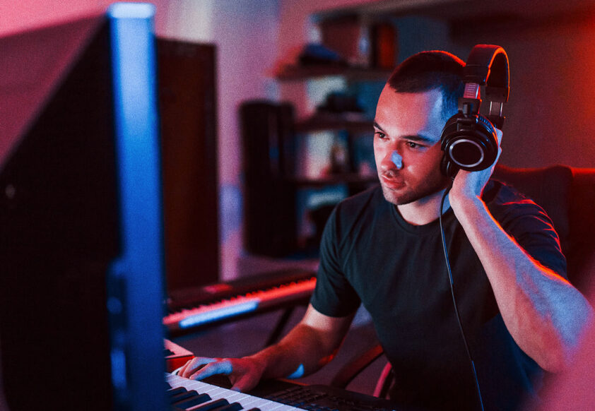 Man sits at keyboard and musical keyboard with headphone to one ear and looks at a computer screen
