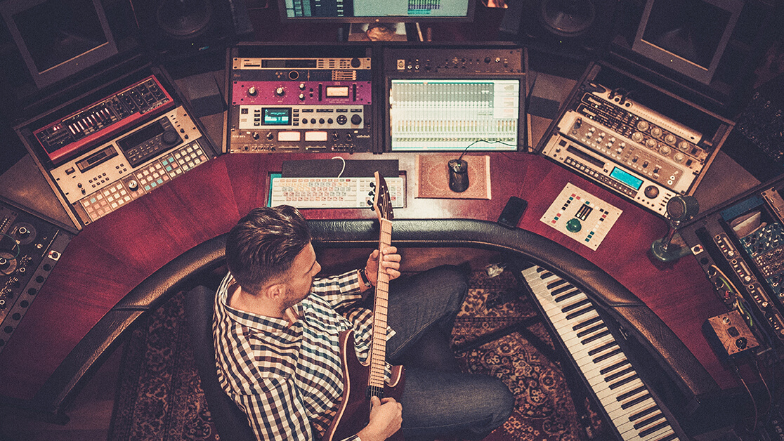 Musician and recording sound engineer sitting at desk with electric guitar in hand. Surrounded by keyboards, mixing consoles and a computer