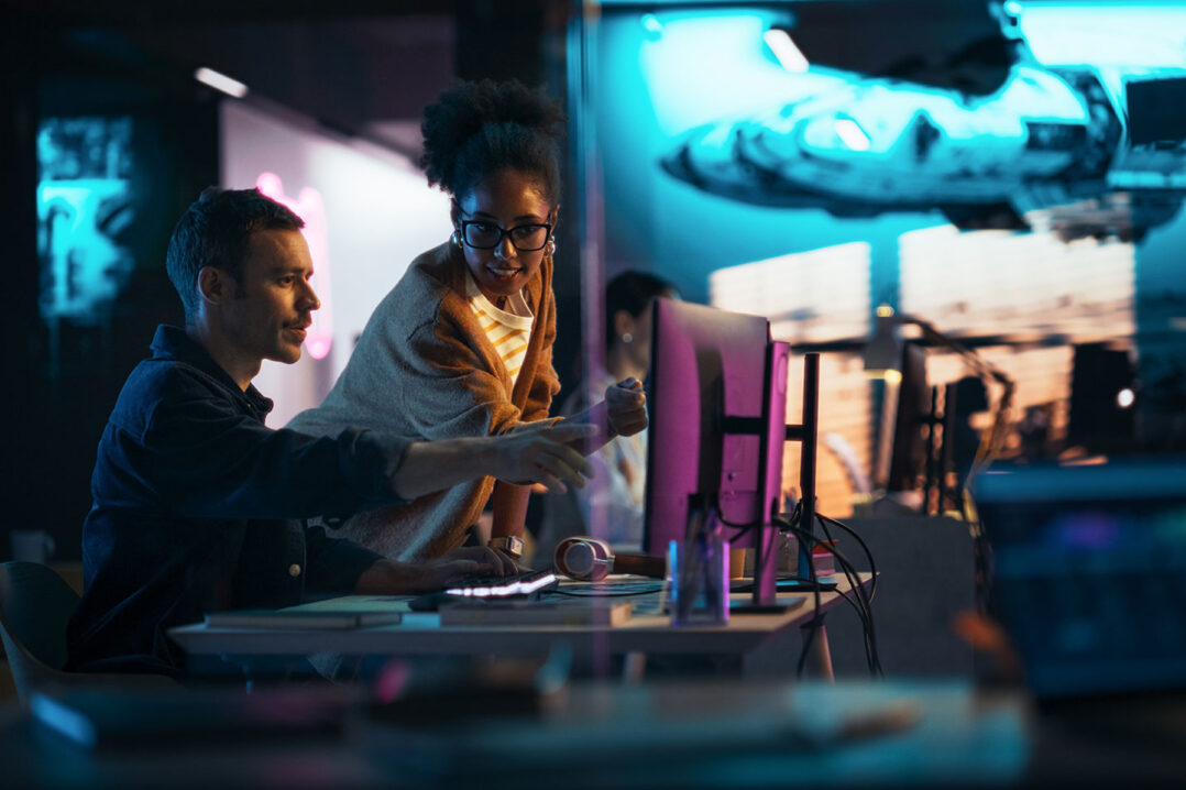 A man sitting and woman standing at a desk in a backlit room, pointing towards a computer monitor