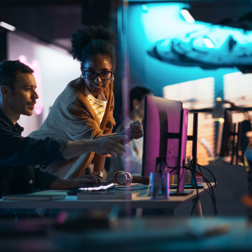 A man sitting and woman standing at a desk in a backlit room, pointing towards a computer monitor