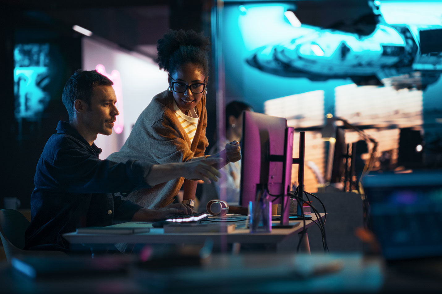 A man sitting and woman standing at a desk in a backlit room, pointing towards a computer monitor