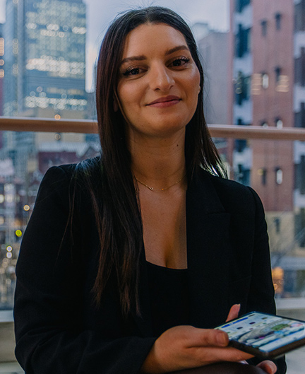 SAE Games Development alumni Rachel Toomeh sitting in front of a window with a mobile phone in her hand and office buildings in the background - mobile image