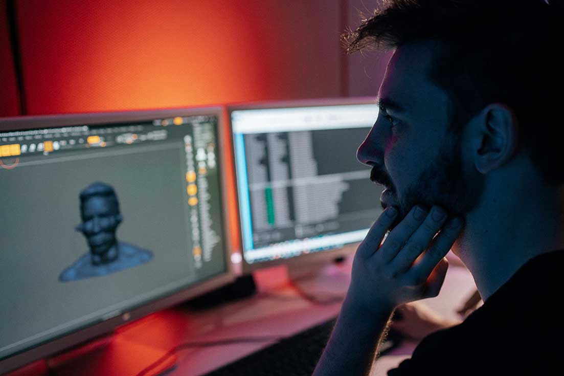 Man sits at two computer screens with Animation modelling software and editing a 3D model