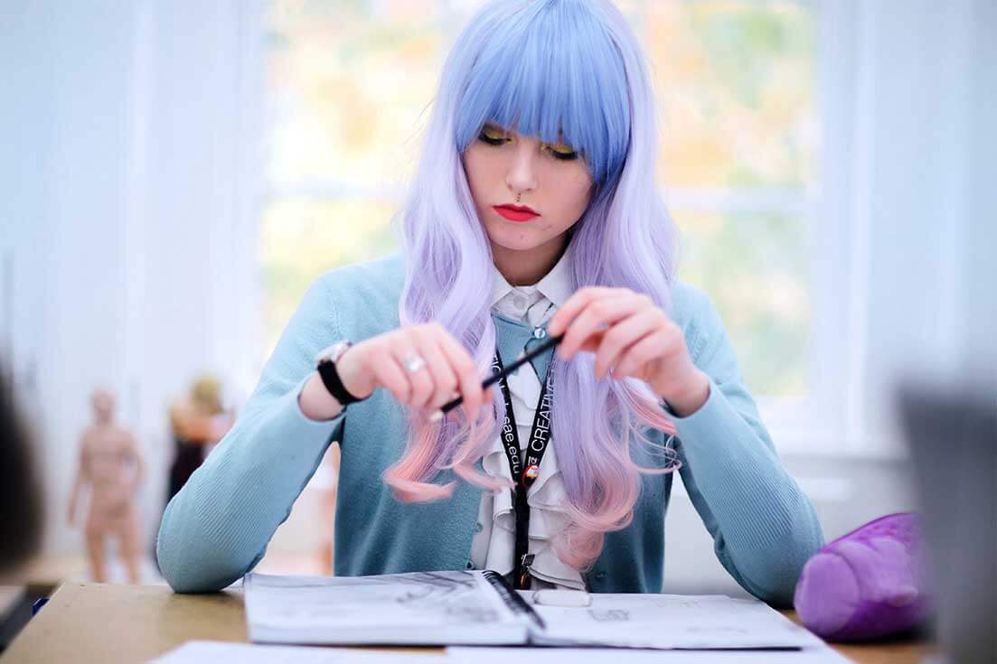 Girl with purple hair sits with pen poised above a sketchpad. SAE Design student