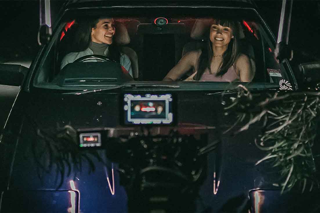 Film set with two females in the front of a car surrounded by cameras, audio and lighting