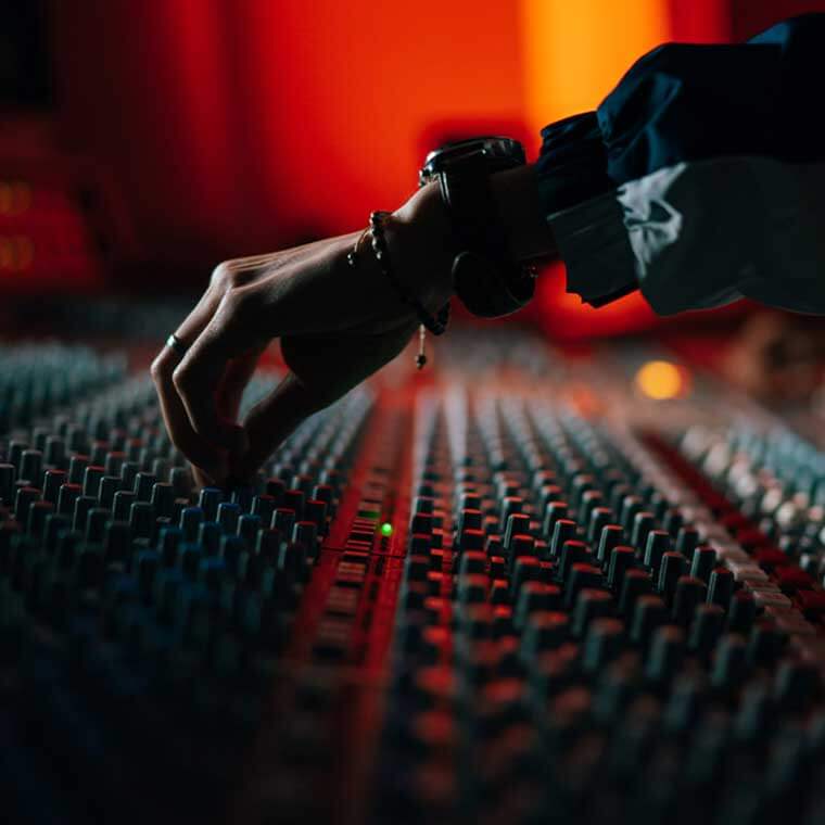 Close up image of a hand and forearm over an analogue mixing desk. Hand is resting on one of hundreds of knobs. Red background light.