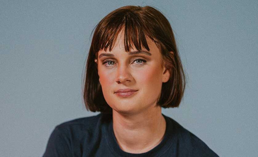 Girl with bobbed red hair and wearing a black tshirt headshot. She is smiling slightly looking straight to camera. SAE alumni and designer Eliza Allard.