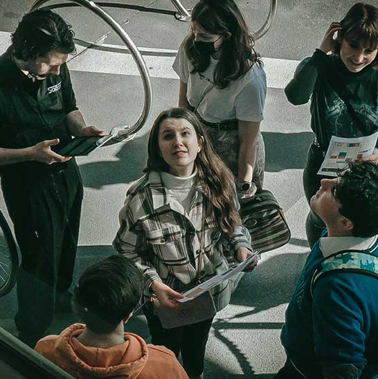 Student holds pamphlet and looks up. A number of people are standing together looking in different directions. Another person holds a tablet and is looking at the screen.