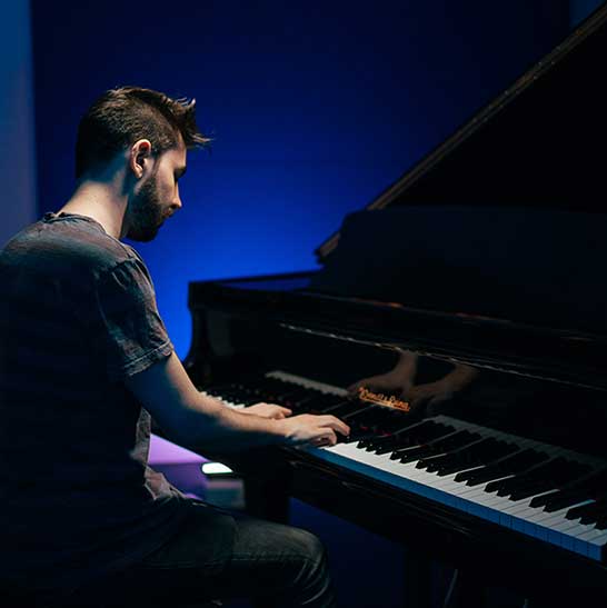Man sits at grand piano with back to us. The piano black and white keys are visible. Blue light against wall behind.
