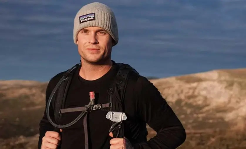 Man wears a beanie and hydration backpack with drinking tube attached to chest. He stands in outdoor location