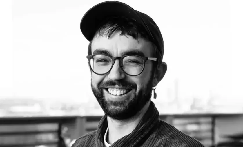 Person with glasses and earring wearing a hat smiles to camera.