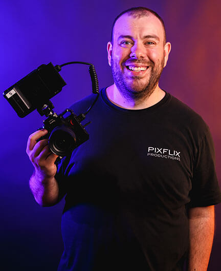 Man wearing a black t-shirt holds camera with viewfinder. The logo or text on the shirt reads Pixflix Productions.