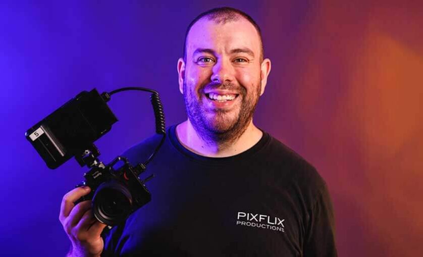 Man with camera wears a t-shirt. Logo on shirt reads: Pixflix Productions
