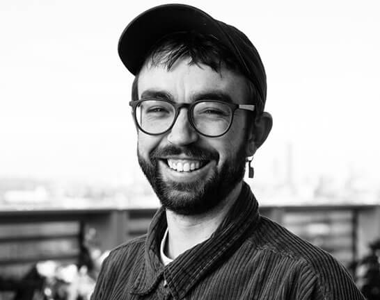 Person smiles at camera. Wears cap and reading glasses. SAE graduate and designer Thomas Hartley