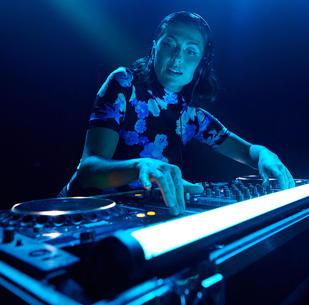 Female DJ mixing music on stage, wearing headphones, with blue lights shining on her and LED light at the front of mixing desk