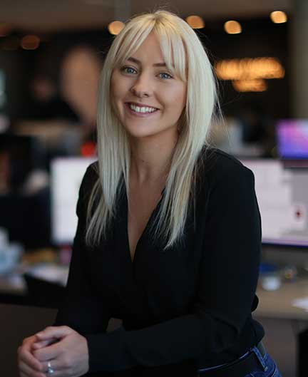 Blonde girl smiles at camera. Wears black shirt and jeans. Her portrait image is taken in front of out of focus office computer screens.