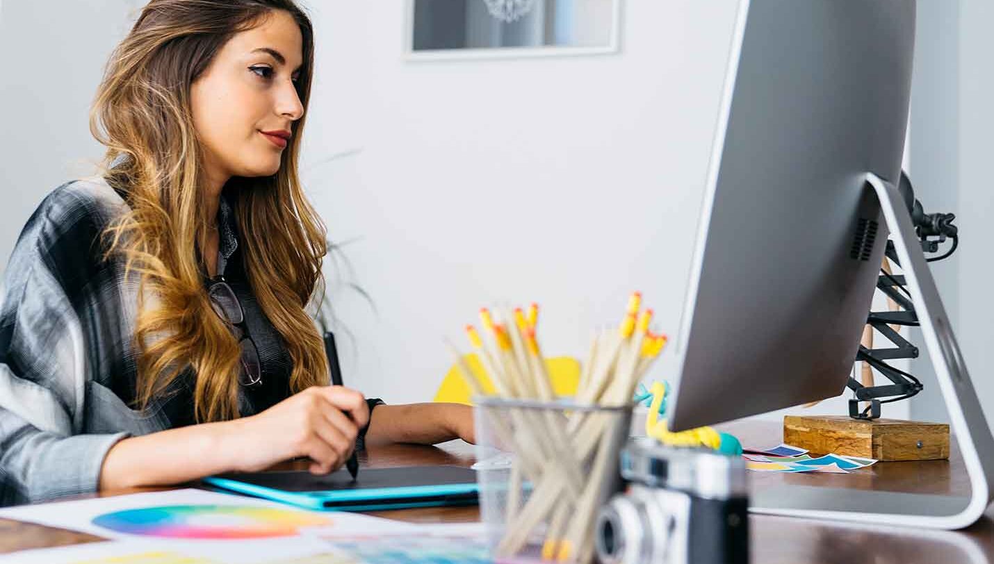 A woman facing side-on to camera at a work desk, looking at a large iMac monitor with a pen in her hand and a colour wheel on her desk | Visual Communication and Graphic Design Courses