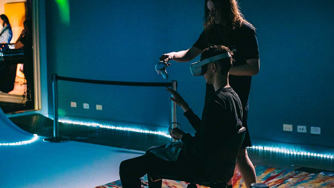 Person sits in a chair with a VR headset and controllers. Person stands and assists person.