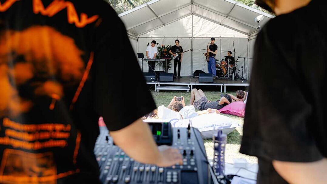 Audio mixing console in foreground. People reclining in the mid ground in front of stage. Four piece band and performing on stage in the background.