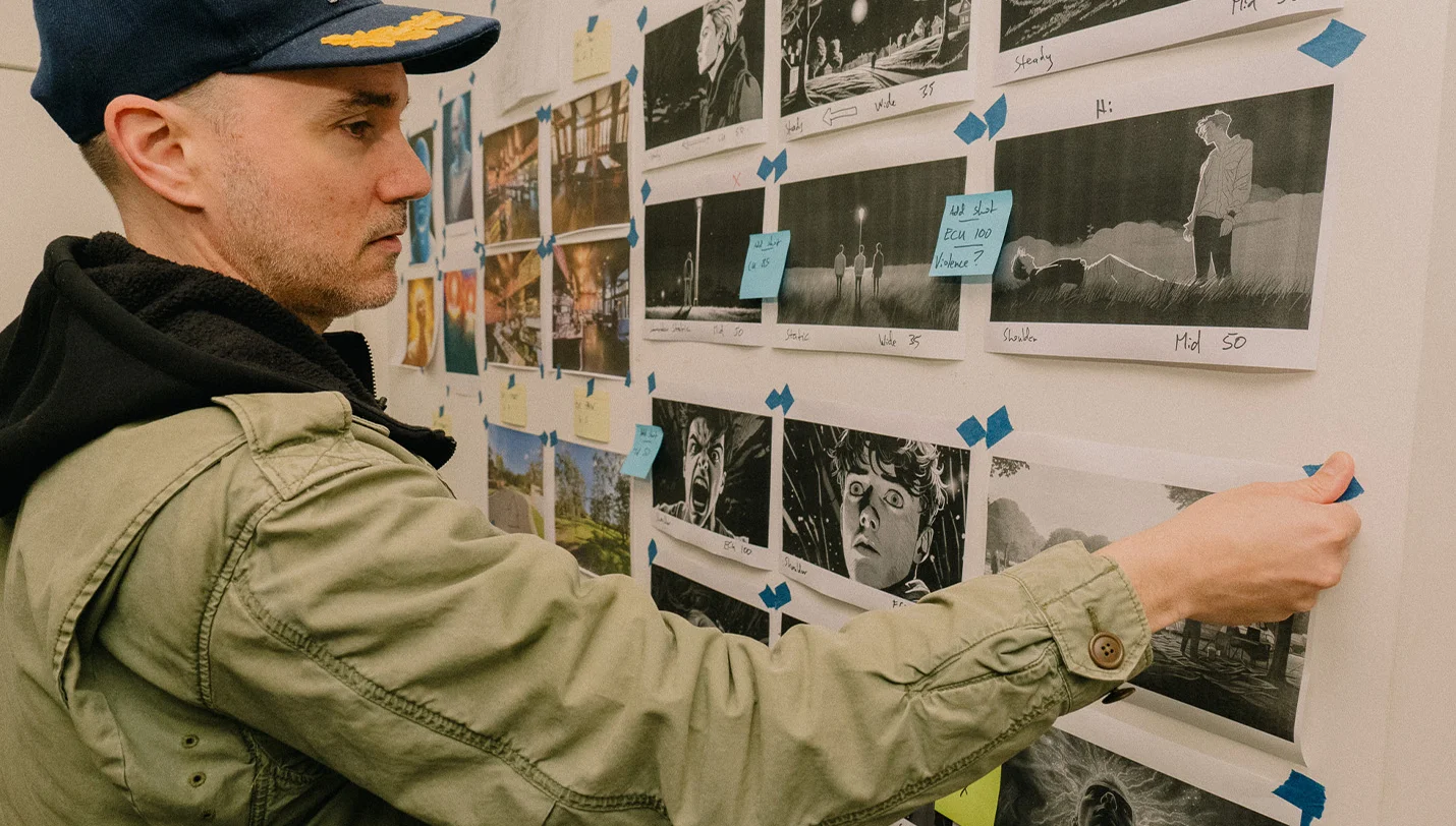 A man wearing a cap stands in front of a wall of animated stills - pictures stuck to the wall. The storyboard of a film. He is pressing the corner piece of tape on one piece of paper.